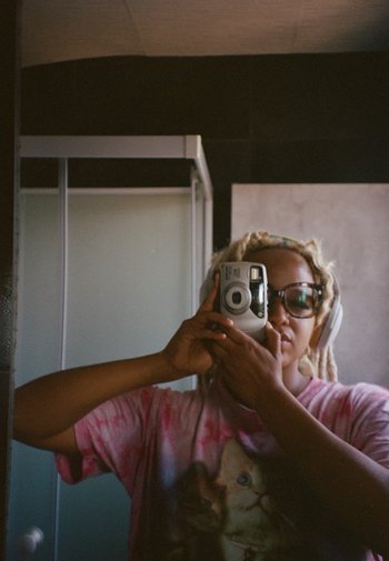 Instantcamera: wit, compact ontwerp. Persoon draagt een bril en koptelefoon, in een roze tie-dye shirt met een kattenafbeelding, staand in een badkamer.