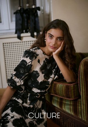 Blumenkleid in Schwarz und Creme mit Puffärmeln und tailliertem Schnitt, kombiniert mit einem schwarzen Gürtel. Sanfte, lockige Haare umrahmen das Gesicht.