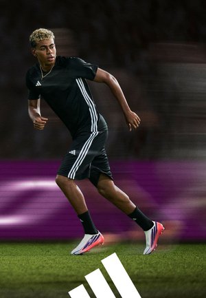 Joven jugador de fútbol masculino con ropa negra de Adidas corriendo en un campo de césped con calcetas negras y botas blancas con suelas rosas.
