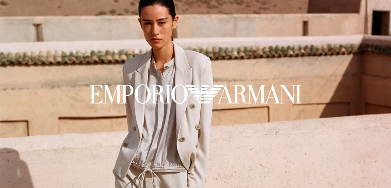 Mujer con traje beige claro y pantalones con cordón, de pie junto a una pared de piedra texturizada en un entorno arquitectónico exterior beige.