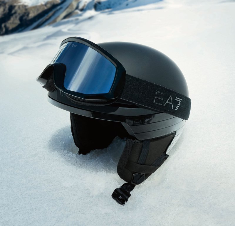 Schwarzer Skihelm mit glatter Oberfläche, verstellbarem Kinnriemen und dunklem Visier. Mit EA7-Logo auf dem Riemen, liegend auf Schnee.