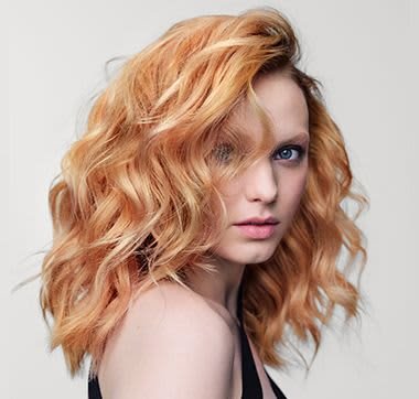 Wavy hair in a light orange shade, textured with volume, framing the face. Neutral background highlights the hair's color and form.