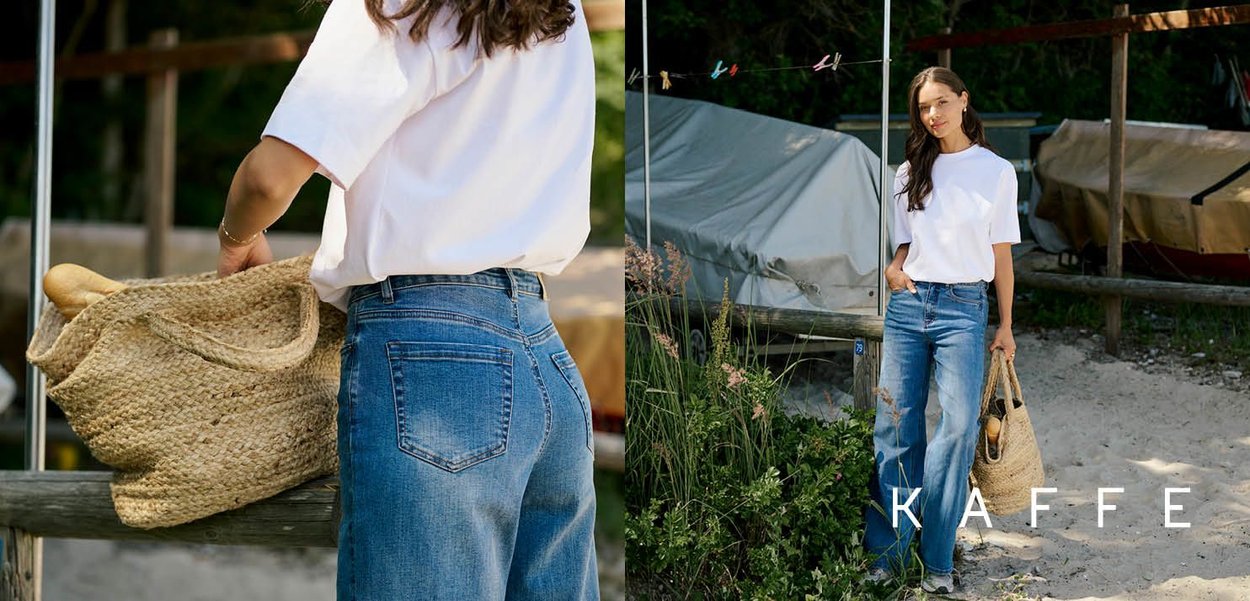 Chemise blanche à manches courtes, jeans en denim bleu taille haute avec une finition décolorée, tenant un sac tissé beige, se tenant sur un sol sablonneux près de bateaux.