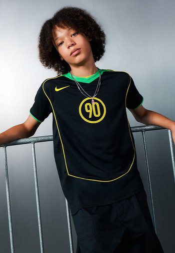 Schwarzes Sporttrikot mit grünen Akzenten und gelber Paspelierung, das ein rundes "90"-Grafikdesign zeigt. Das Modell trägt übereinanderliegende Halsketten und schwarze Shorts.