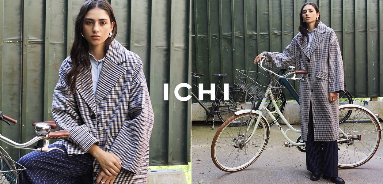 Cappotto lungo a scacchi con ampio colletto, tessuto strutturato e grandi tasche, indossato su pantaloni a righe. Bicicletta sullo sfondo, ambiente urbano.