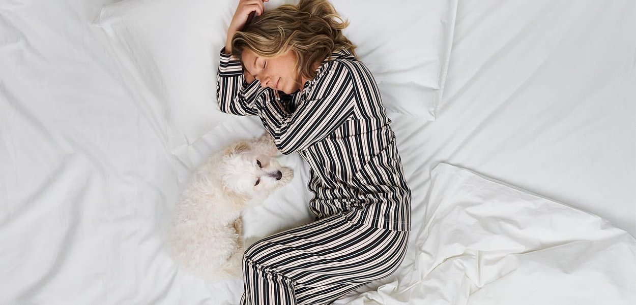 Striped pajama set in black, cream, and rust, with a soft texture, worn by a person lying on white sheets beside a small white dog.