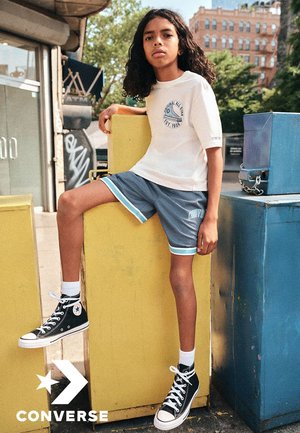 Zapatillas altas Converse negras, pantalones cortos deportivos grises con acentos en azul aguamarina y una camiseta blanca con un logo circular. De pie frente a contenedores coloridos.