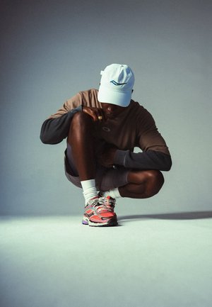 Tenue de sport avec un haut à manches longues marron, un short gris, des chaussettes blanches, et des chaussures de course colorées avec un motif en zigzag. Casquette blanche portée.