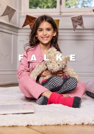 Child wearing a pink coat, holding a teddy bear. Features red and black polka dot socks. Set on a soft rug in a light interior.
