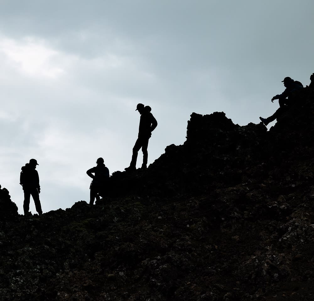 Silhouette di cinque escursionisti su un terreno roccioso, che indossano cappelli e si trovano in piedi o seduti. Cielo nuvoloso sullo sfondo. Texture scura delle rocce visibile.