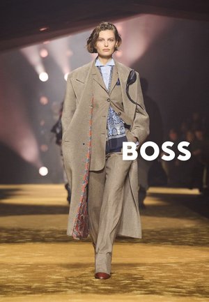 Vrouwelijk model loopt over de catwalk met een oversized beige pak met een patroon aan de binnenkant en een blauw overhemd, verlicht door catwalk-spots, met de tekst BOSS eroverheen.