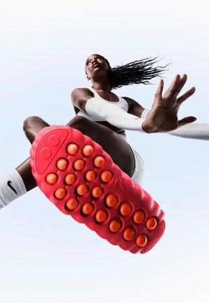 Czerwony but sportowy z teksturowaną podeszwą wyposażoną w duże pomarańczowe okrągłe wypustki, przedstawiony w powietrzu na jasnoniebieskim tle.