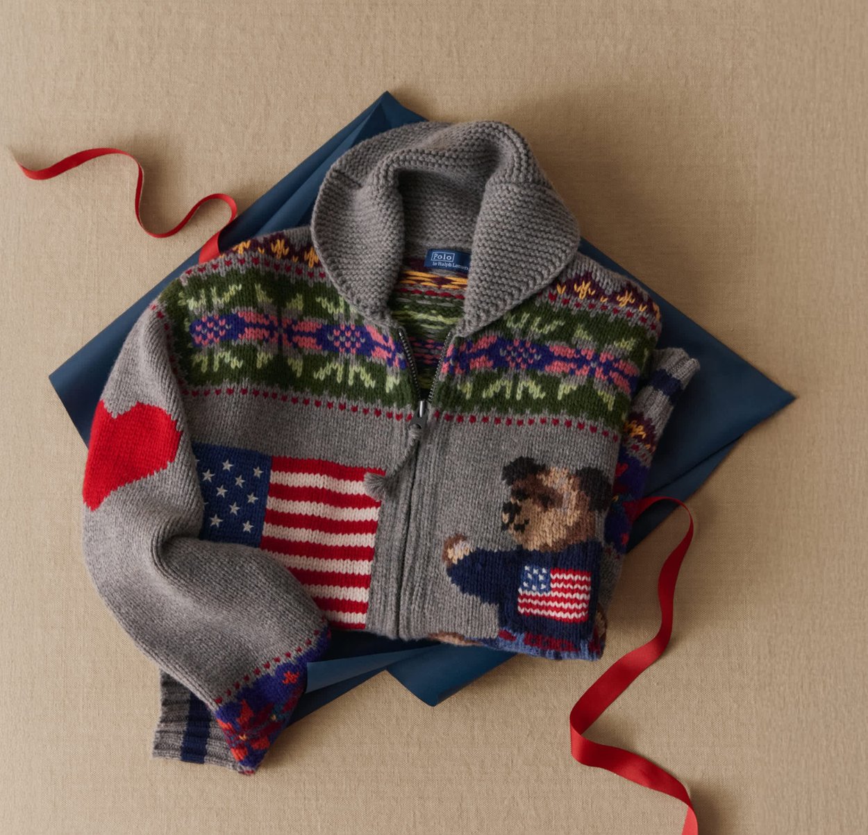 Pull gris zippé avec des motifs colorés, y compris un ours et des accents du drapeau américain, en maille texturée avec manches repliées.