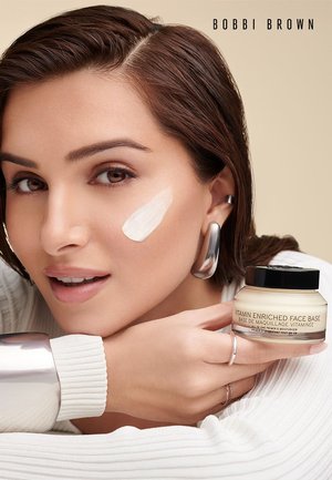 Femme aux cheveux brun clair et aux bijoux en argent applique une crème blanche sur sa joue, tenant un pot étiqueté "Base de Visage Enrichie en Vitamines" de Bobbi Brown.