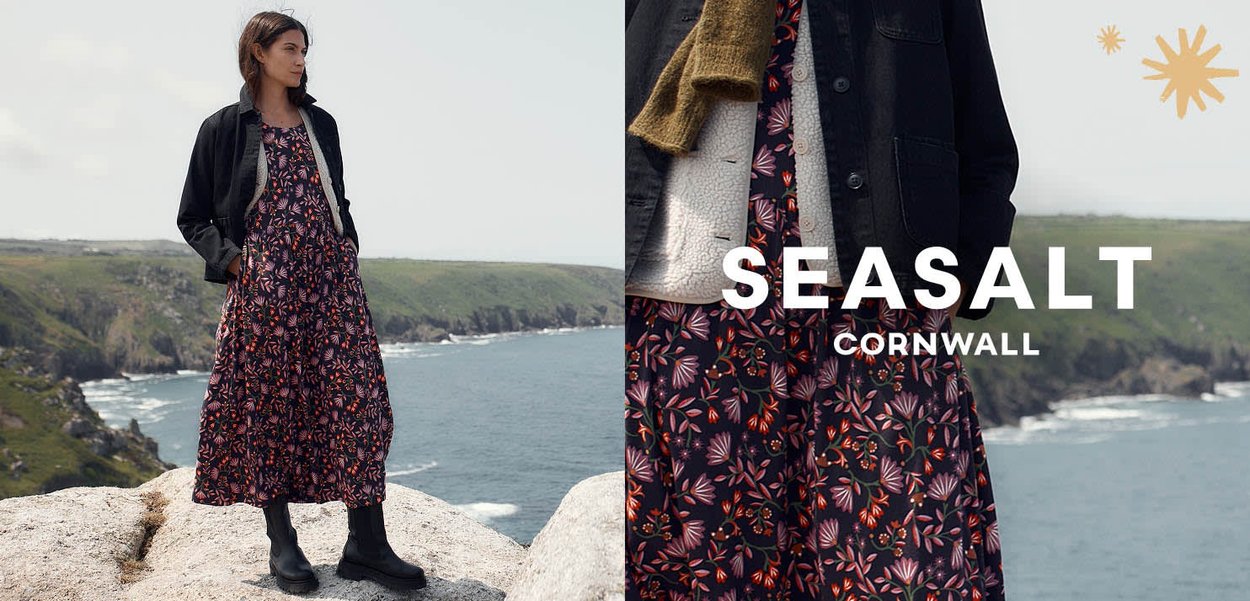 Maxikleid mit Blumenmuster in dunklen Tönen, kombiniert mit einer schwarzen Jacke und einem beigefarbenen Pullover. Dazu schwarze Stiefeletten, vor einer Küstenlandschaft.