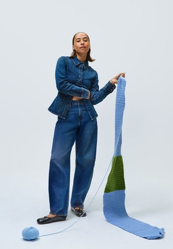 Denimjack en wijd-uitlopende jeans in donkerblauw, met een veelkleurig gebreid stuk met blauwe en groene secties, met een blauwe bol garen op de grond.