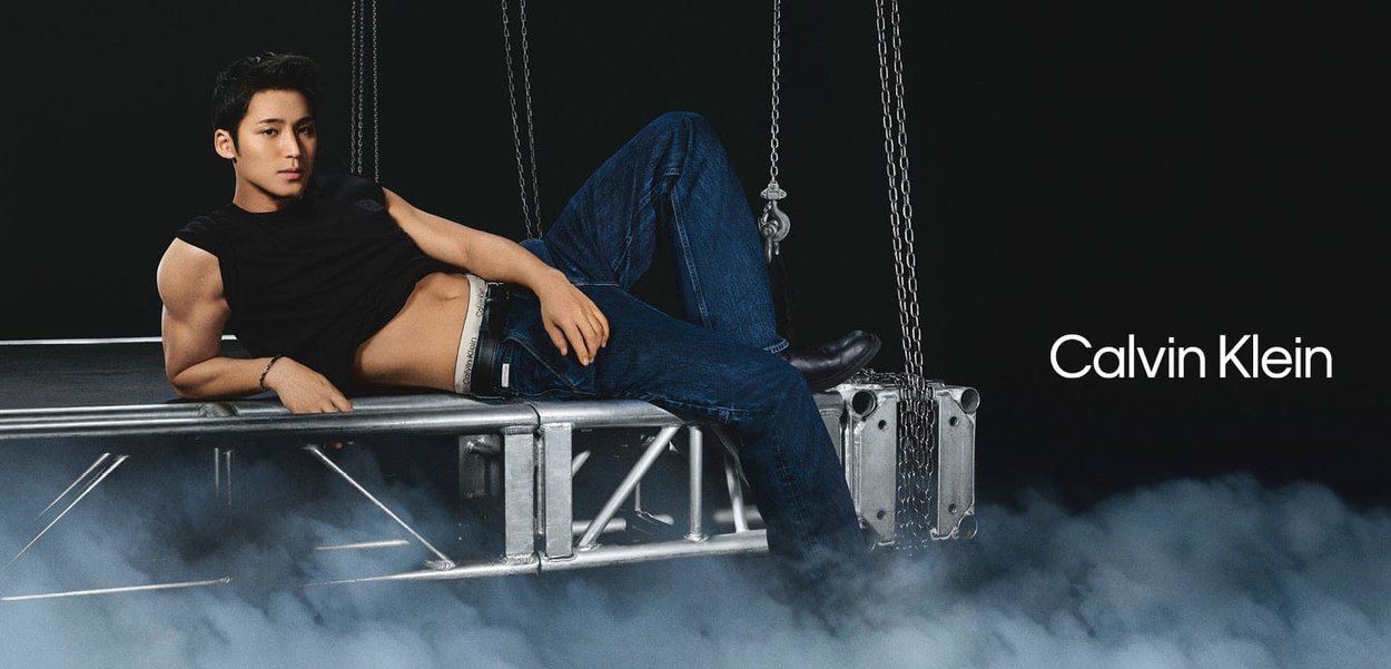 Man wearing black sleeveless top and dark blue jeans reclines on metal platform suspended by chains, with smoke below and Calvin Klein logo.