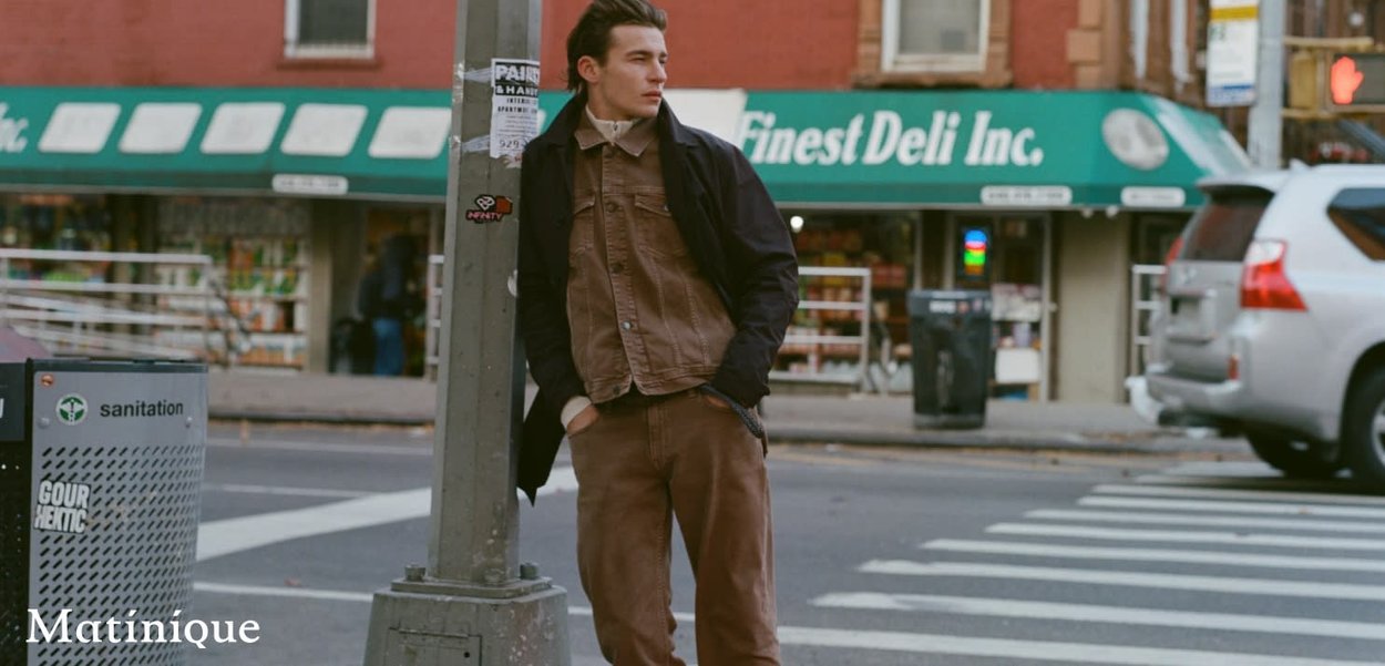 Junger Mann in brauner Jacke und Hose lehnt an einem Straßenlaternenpfahl nahe eines Feinkostgeschäfts an einer städtischen Kreuzung.