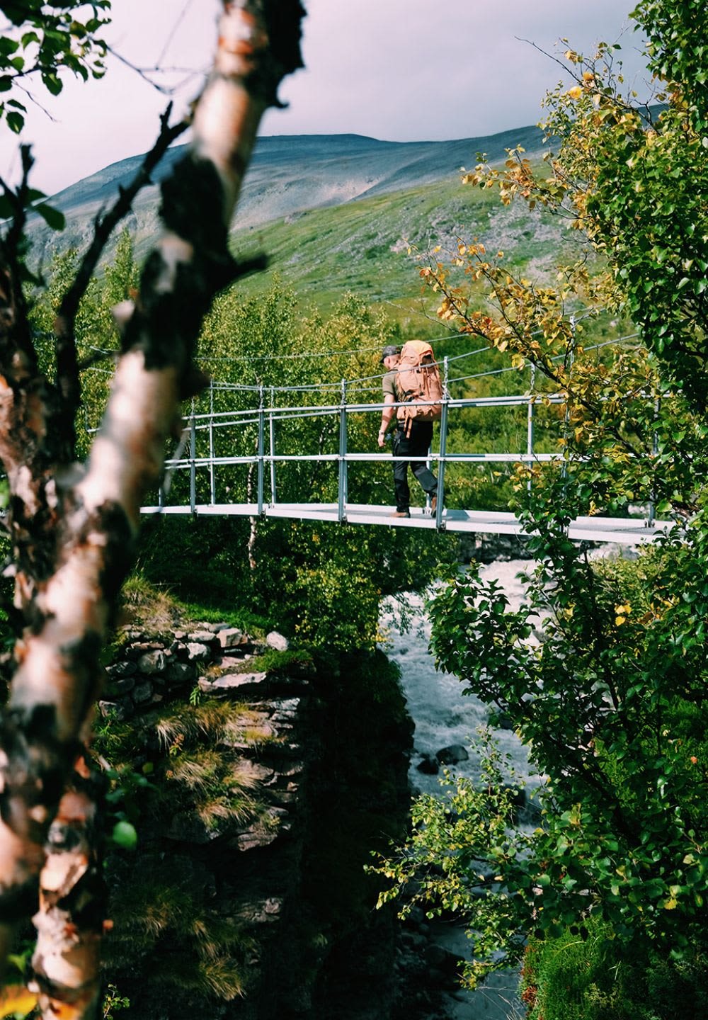 Bro av metall med en enkel design. En person går över den med en brun ryggsäck, omgiven av grönska och en flödande flod nedanför.