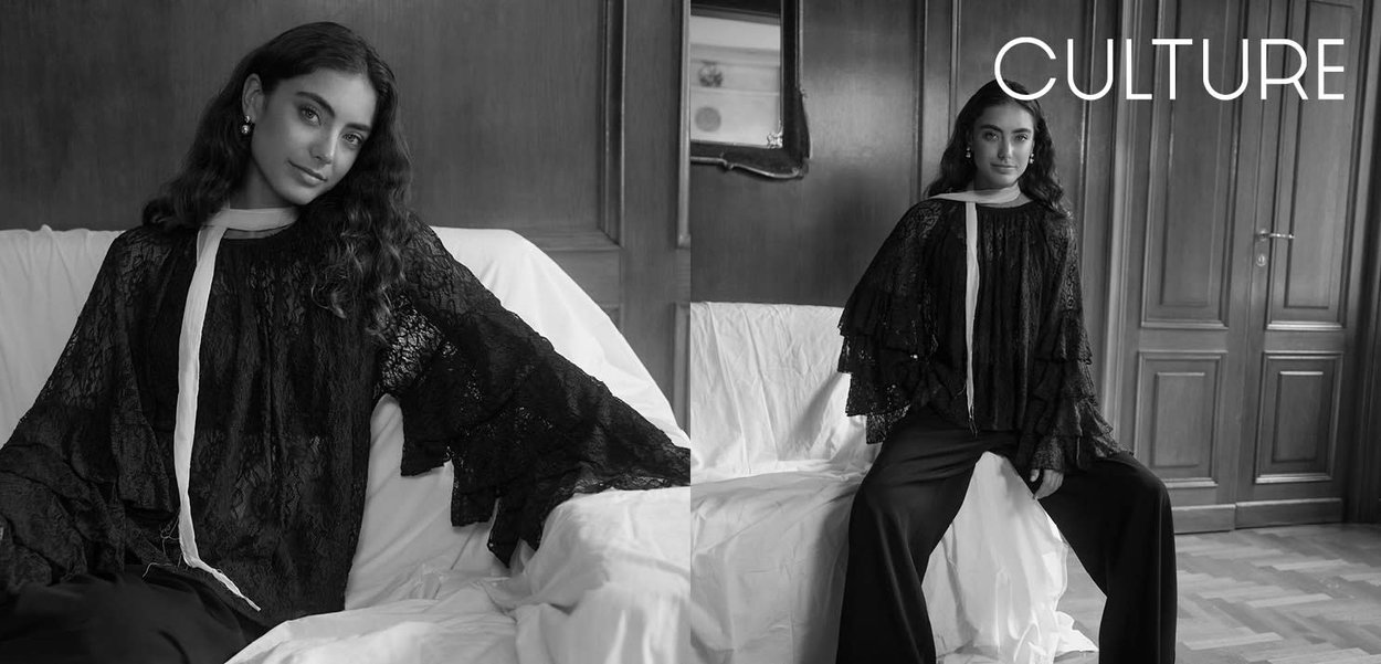 Schwarze Spitzenbluse mit gerafften Ärmeln, weißem Halstuch-Detail; kombiniert mit schwarzer Hose. Im Hintergrund sind Holzpaneele und ein Sofa zu sehen.