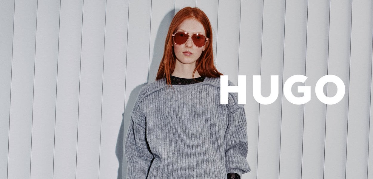 Pull gris tricoté avec manches amples, associé à des lunettes de soleil rondes à verres rouges. Arrière-plan avec des stores verticaux. Texte "HUGO" à droite.