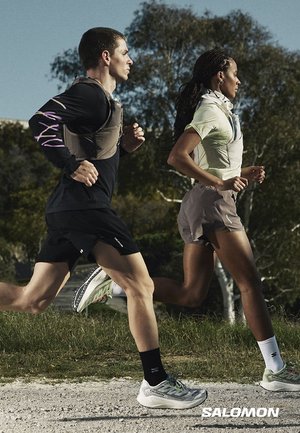 Due corridori, uno uomo e una donna, indossano abbigliamento sportivo e zaini idrici, correndo all'aperto su un sentiero ghiaioso con alberi sullo sfondo.