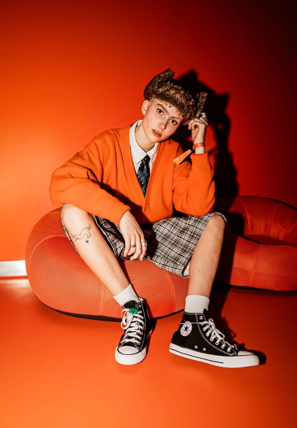 Orange cardigan, white shirt, plaid shorts. Black Converse shoes with white soles and green accents. Sitting on a red rounded sofa.