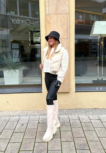Pufferväst i krämfärg, ribbade tröja, svarta leggings och knähöga vita stövlar. Svart bucket hat, stående mot en butiksmiljö.
