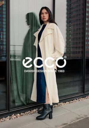Cappotto lungo color crema con rever oversize, indossato sopra un vestito blu. Stivaletti neri alla caviglia con tacco largo. Finestre di vetro sullo sfondo.