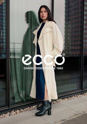 Cappotto oversized crema con ampi risvolti, abbinato a un vestito blu scuro e stivali neri alla caviglia su una superficie testurizzata. Riflesso in uno sfondo di vetro.