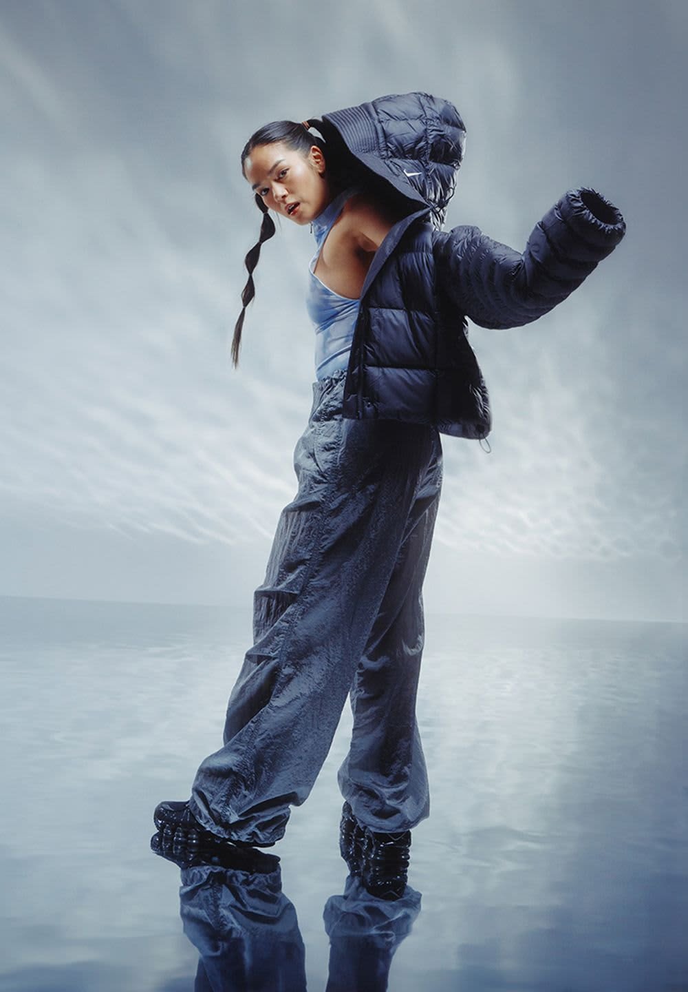 Puffy black jacket with one sleeve, blue tank top, textured gray pants, and glossy black footwear. Smooth reflective surface backdrop.