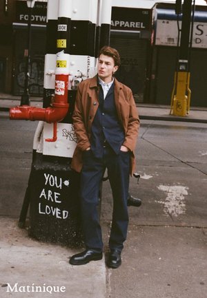 Giovane uomo con cappotto marrone e abito blu scuro appoggiato a un palo bianco e nero coperto di graffiti, con la scritta "SEI AMATO" su un marciapiede cittadino.