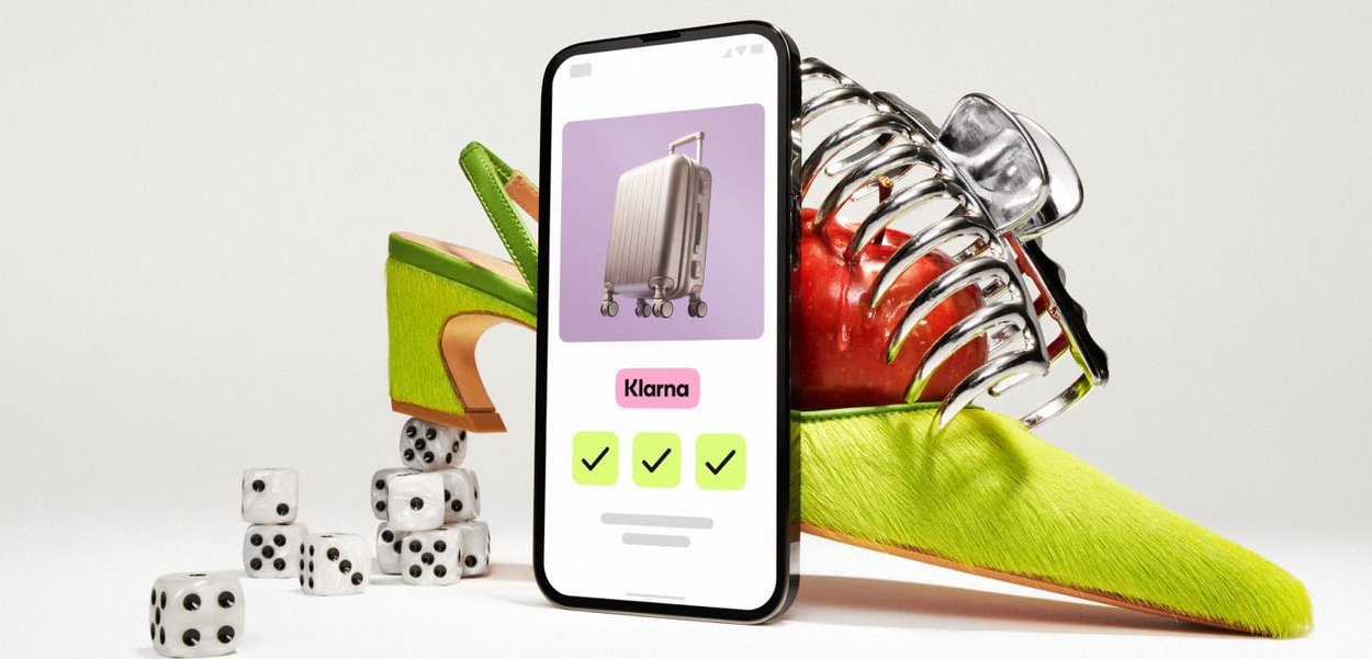 Smartphone, das einen silbernen Koffer vor einem lila Hintergrund zeigt, umgeben von einem grünen High-Heel-Schuh, einem Apfel und Würfeln.