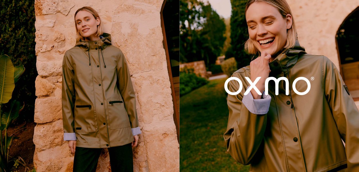 Kvinde iført en grøn regnjakke står og smiler udendørs nær en stenmur og en have, hvor hun viser en afslappet vandtæt jakke fra Oxmo.