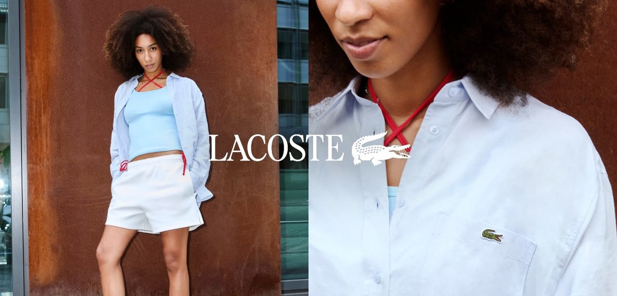 Donna con capelli ricci che indossa una camicia azzurra, pantaloncini bianchi e top blu, in piedi contro un muro color ruggine con il logo del marchio visibile.