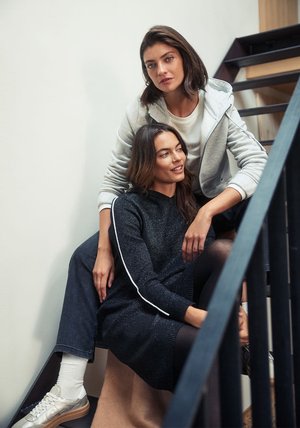 Dwie kobiety pozują na schodach. Jedna ma na sobie szarą bluzę z kapturem na białym topie; druga nosi ciemną, błyszczącą sukienkę z białymi paskami.