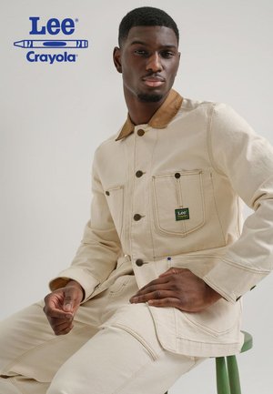 Man wearing a cream-coloured jacket and trousers from the Lee and Crayola collaboration, seated on a green stool against a neutral background.
