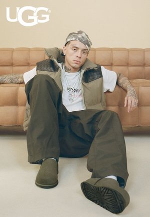 Jeune homme tatoué portant un bandana, des vêtements en couches et des chaussures slip-on UGG vertes, assis par terre devant un canapé beige.