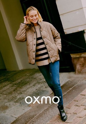Beige quiltet jakke med trykknapper, båret over en sort og beige stribet sweater, parret med blå smalle jeans og sorte ankelstøvler.