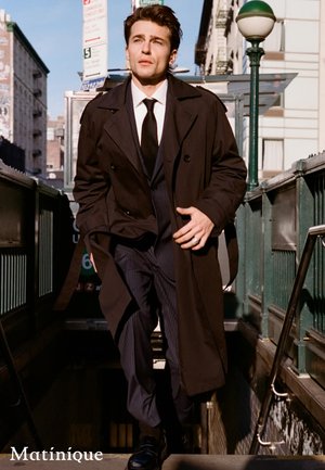 Mann in schwarzem Trenchcoat und Anzug, der in der Stadt eine U-Bahn-Treppe hinaufgeht, Sonnenlicht wirft Schatten auf ihn und die umliegenden Gebäude.