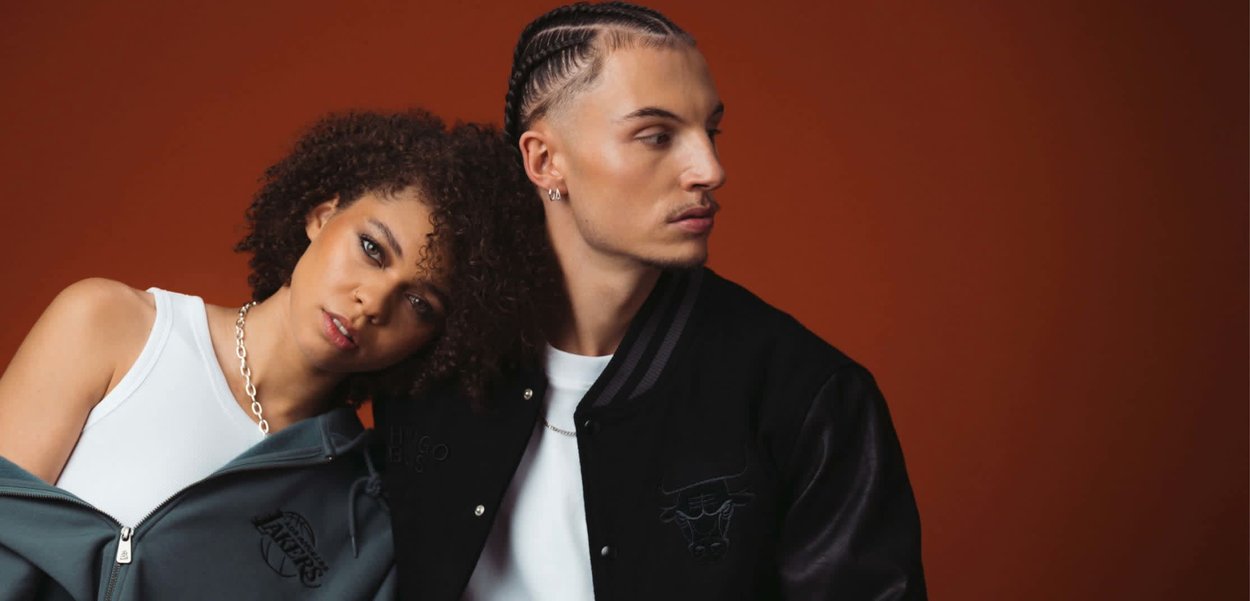 Donna con capelli ricci appoggia la testa sulla spalla di un uomo con capelli intrecciati; entrambi indossano giacche casual su uno sfondo marrone.