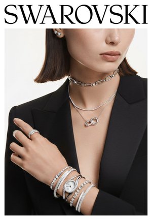 Biżuteria Swarovski z wisiorkiem w kolorze srebra, naszyjnikami warstwowymi oraz wieloma bransoletkami z błyszczącymi akcentami i zegarkiem. Czarna marynarka w tle.