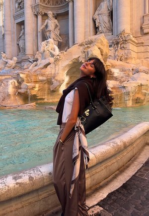 Borsa in pelle nera con accessori in argento, indossata a tracolla. Modella con top bianco e pantaloni marroni a vita alta, vicino a una fontana di pietra.