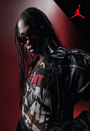 Persona con capelli lunghi intrecciati, occhiali da sole, giacca di pelle nera e T-shirt grafica a tema basket, su sfondo rosso.