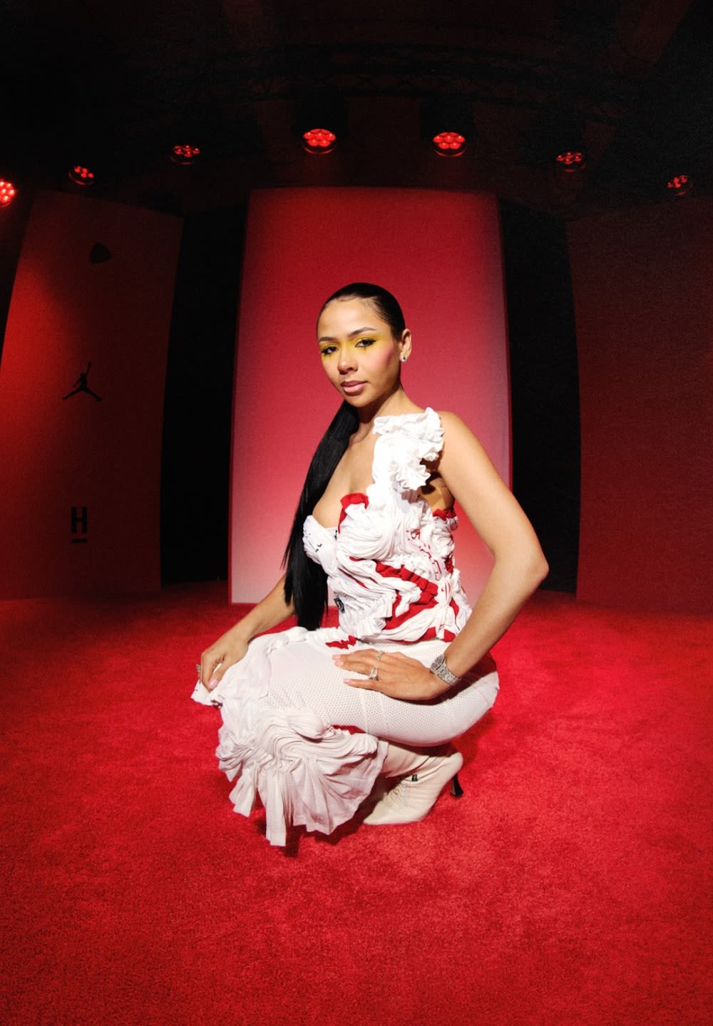 Mujer con cabello largo y oscuro vestida con un vestido blanco y rojo texturizado, arrodillada sobre una alfombra roja bajo luces rojas de escenario.