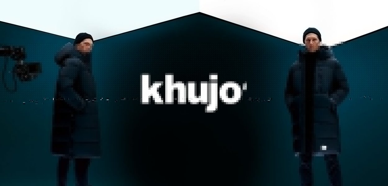 Musta toppatakki, jossa on huppu, sivutaskut ja logoplaatta. Malli seisoo geometrista taustaa vasten kameran kanssa.