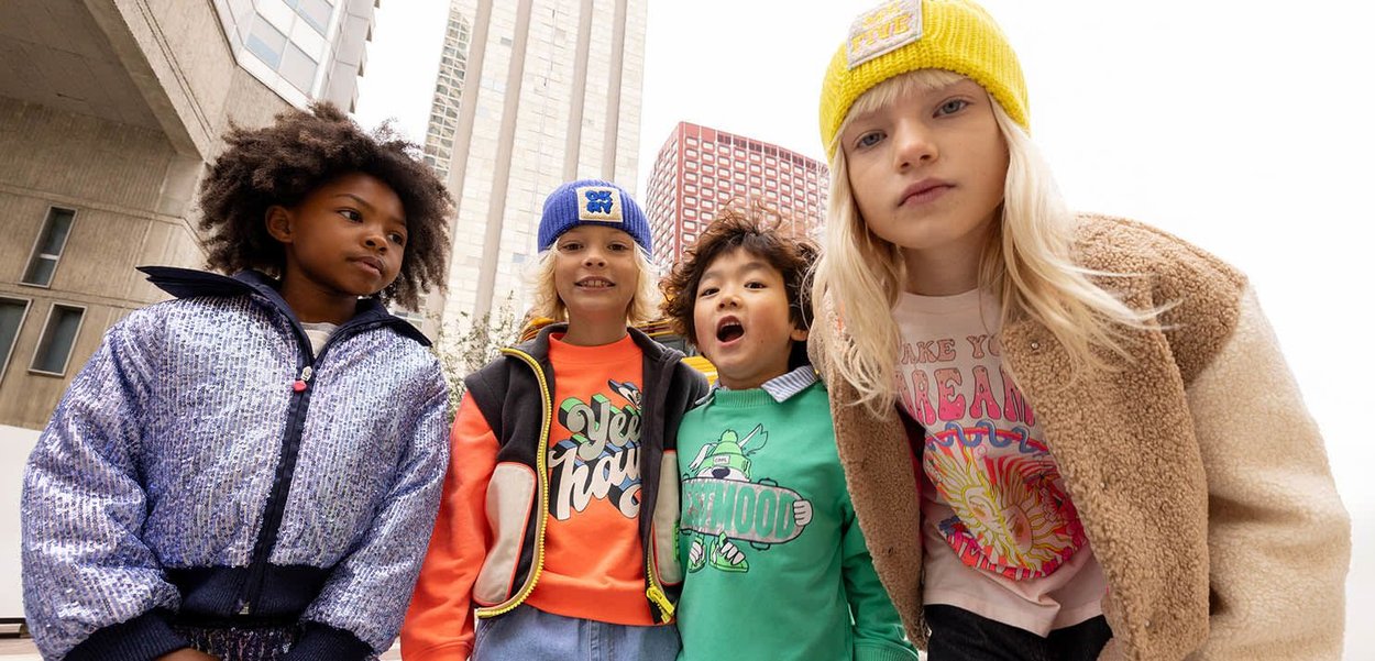 Quatre enfants posent, vêtus d'une veste à sequins, de sweats colorés, d'un pull vert et d'une veste en fausse fourrure beige, avec diverses bonnets en tricot.