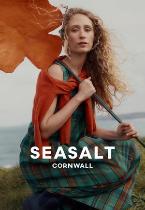 Vrouw met lang krullend haar houdt groot oranje zeewier vast, draagt een groene geruite jurk en een oranje trui, zittend op een grasrijke kust vlak bij zee.