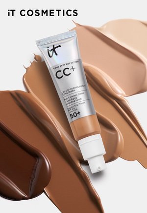 Tube der IT Cosmetics CC+ farbkorrigierenden Sonnencreme mit SPF 50+ vor einem Hintergrund aus verschmierten Foundation-Farbtönen von hell bis dunkel.