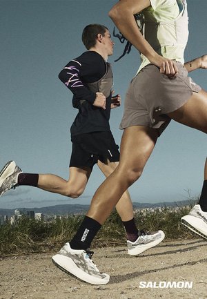 To løbere, en mand og en kvinde, iført sportstøj og Salomon-sko løber på en jordsti med byens skyline i baggrunden.